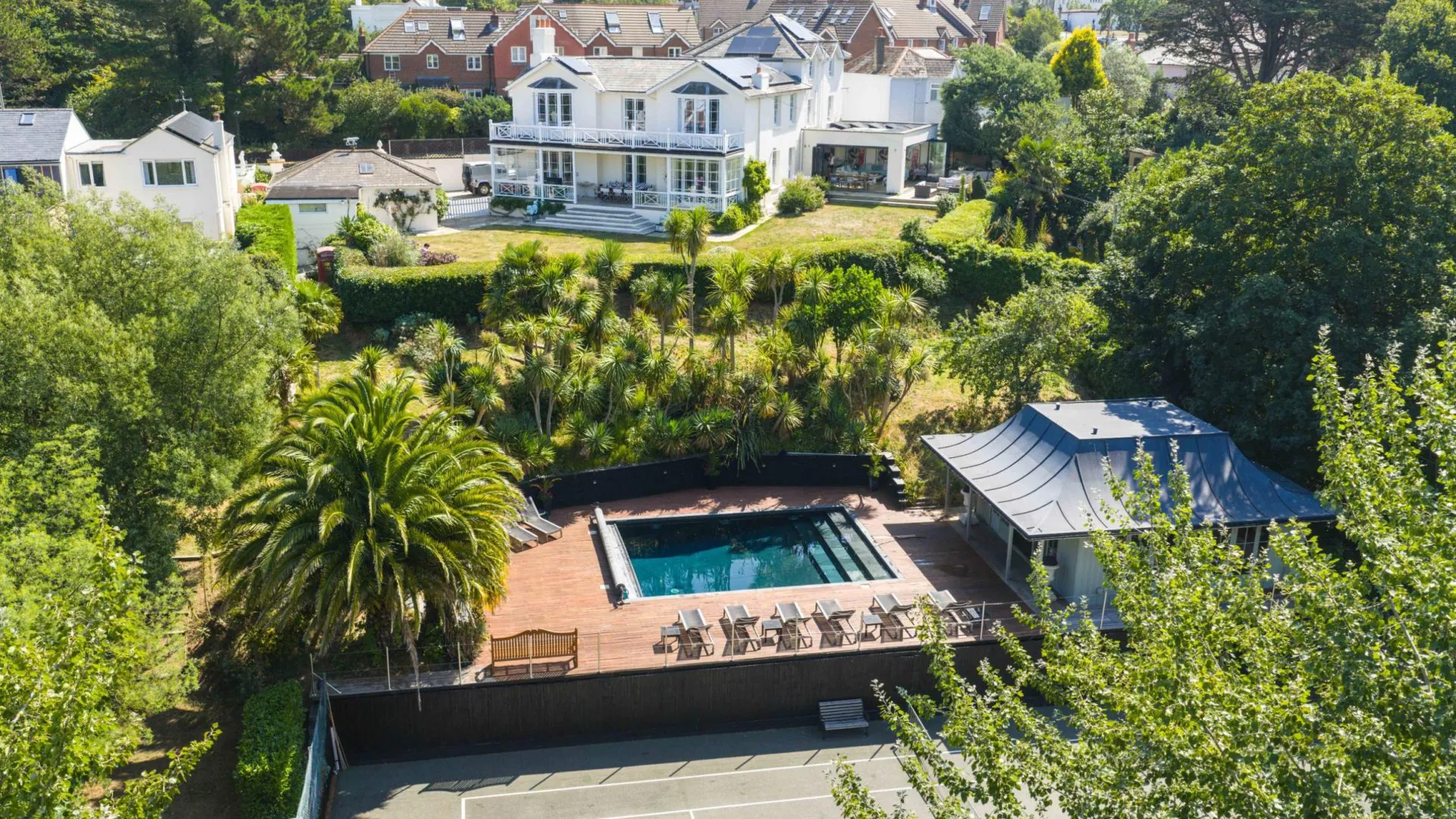 Large white house with balconies overlooking landscaped gardens, a pool terrace with sun loungers, and a tennis court below.