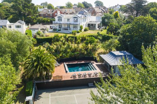 Large white house with balconies overlooking landscaped gardens, a pool terrace with sun loungers, and a tennis court below.